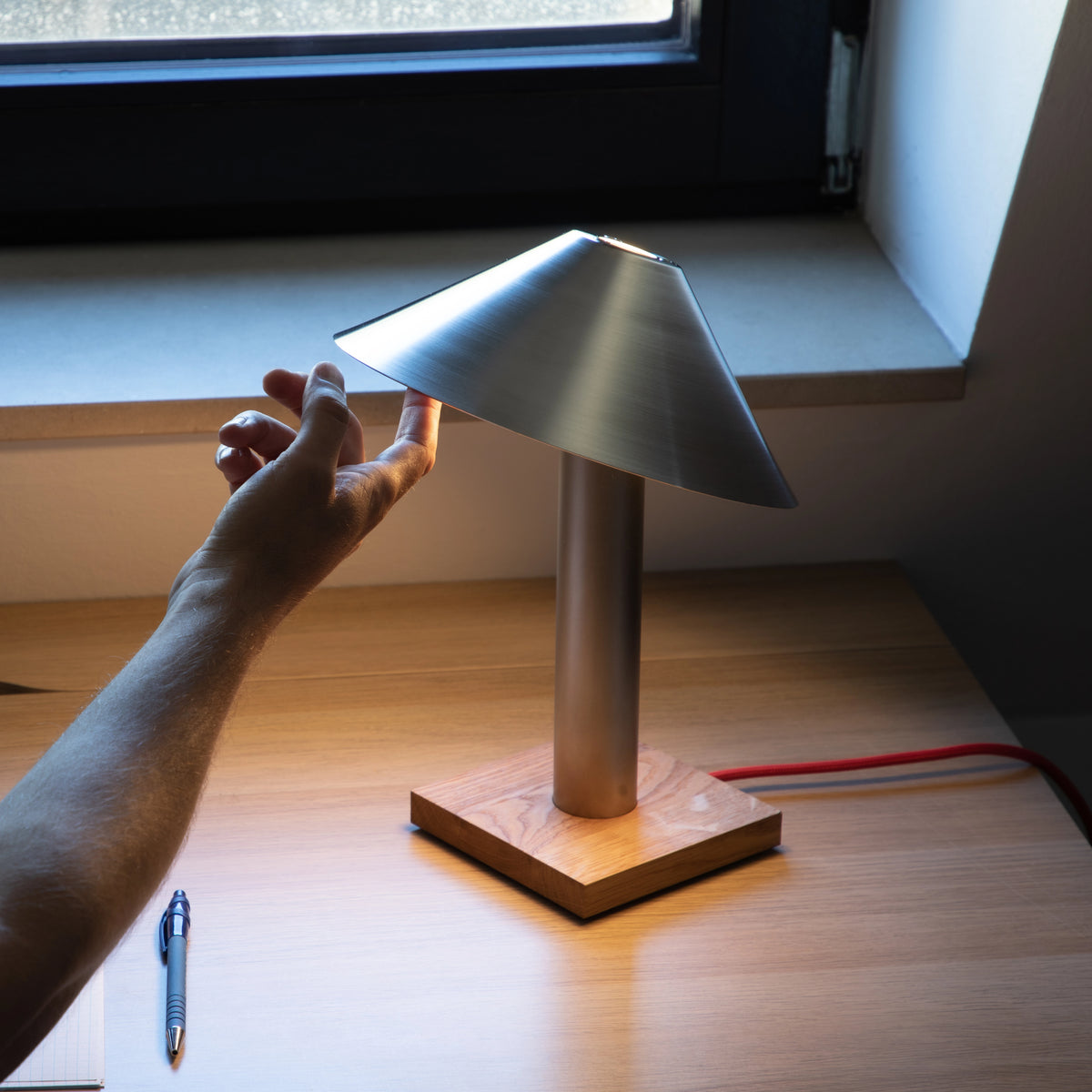 Hand kippt den Edelstahlschirm der Tischleuchte PFEIL im Gegenlicht am Fenster, warmes Licht darunter