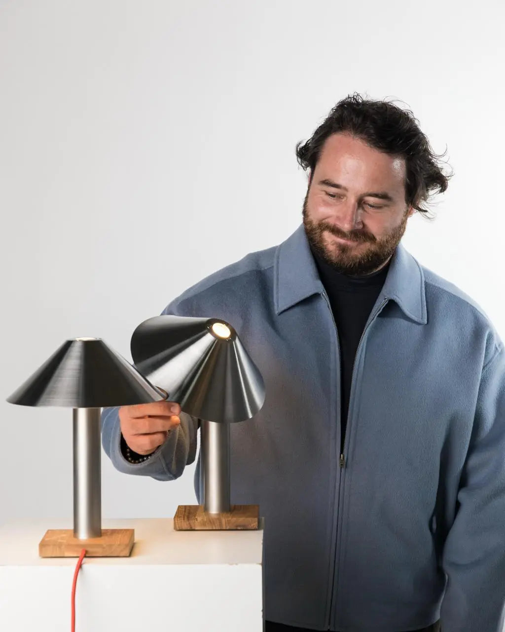 Zwei Tischleuchten PFEIL auf hellem Podest, eine Person kippt den Edelstahlschirm der leuchtenden Lampe