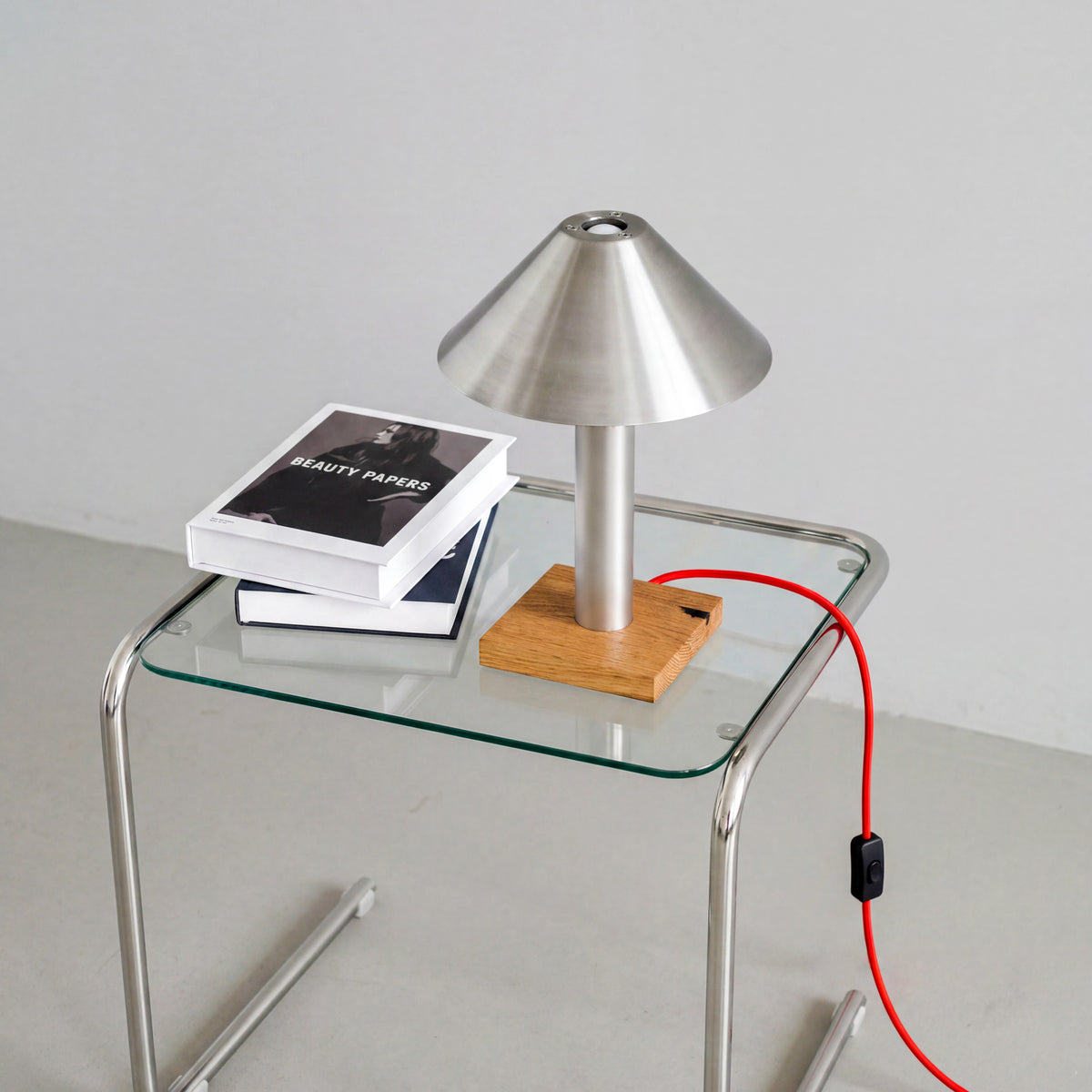 Tischleuchte PFEIL aus Edelstahl und Eiche auf einem Beistelltisch aus Glas und Chrom, mit rotem Textilkabel und Kippschalter.